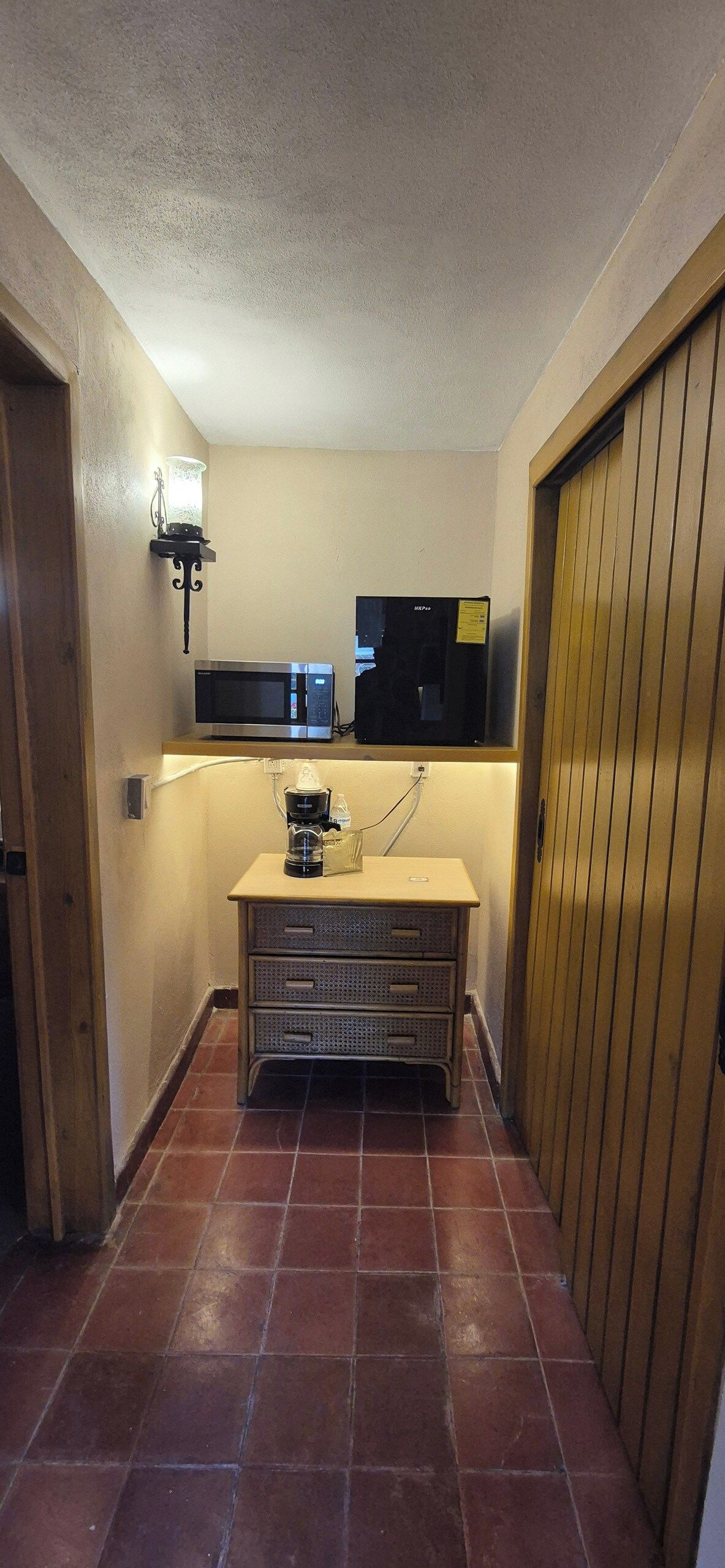A narrow hallway with a microwave and a black appliance on a shelf, a coffee maker on a cabinet, and a wooden door on the right.