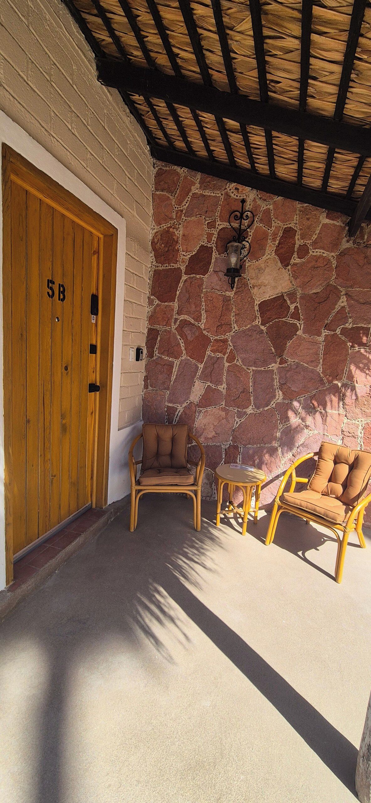 Un muro de piedra con una puerta de madera marcada '5B' y dos sillas de mimbre.