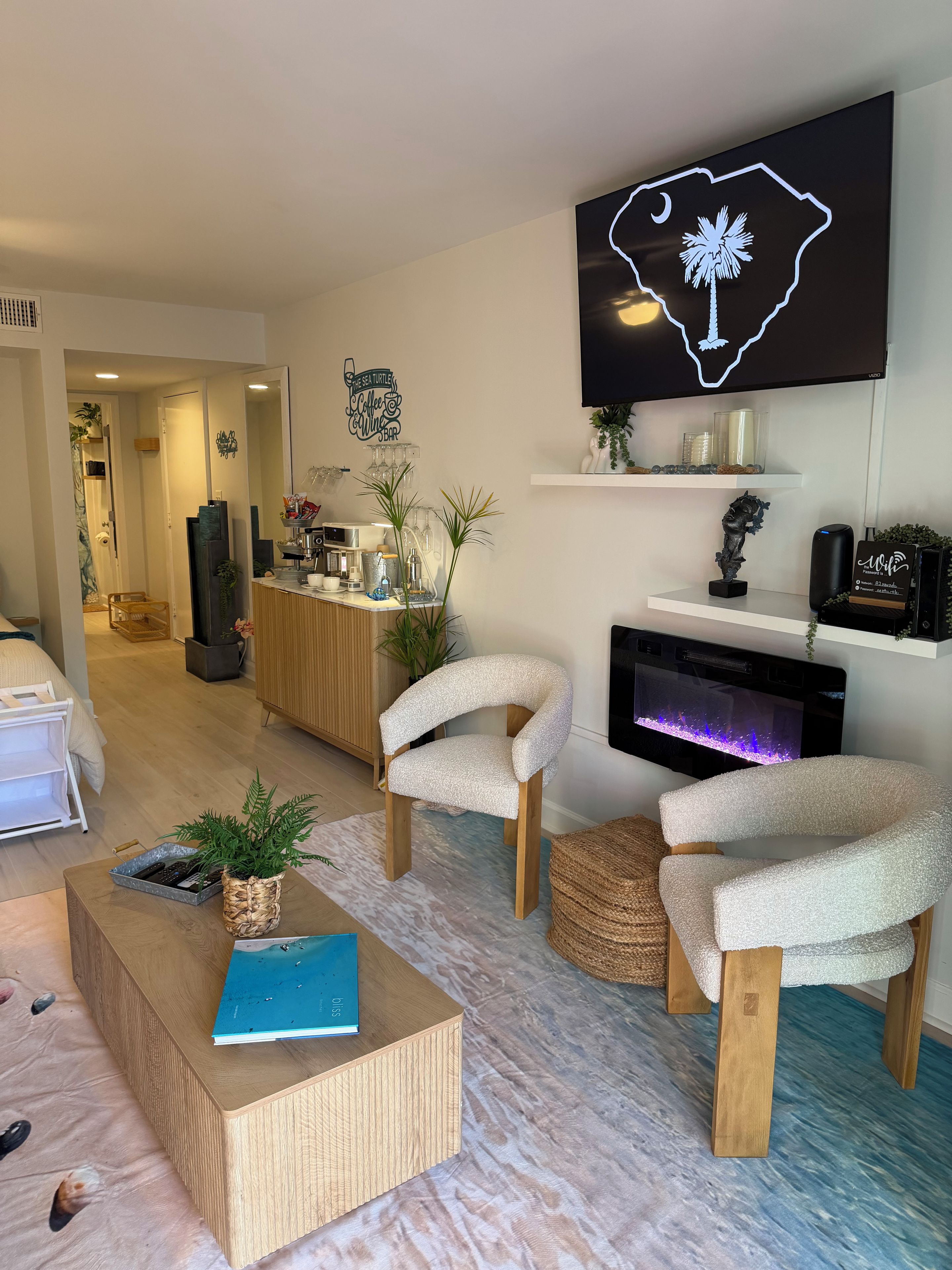 A modern living room with a flat-screen TV, a coffee table, two armchairs, a fireplace, and a kitchen area in the background.