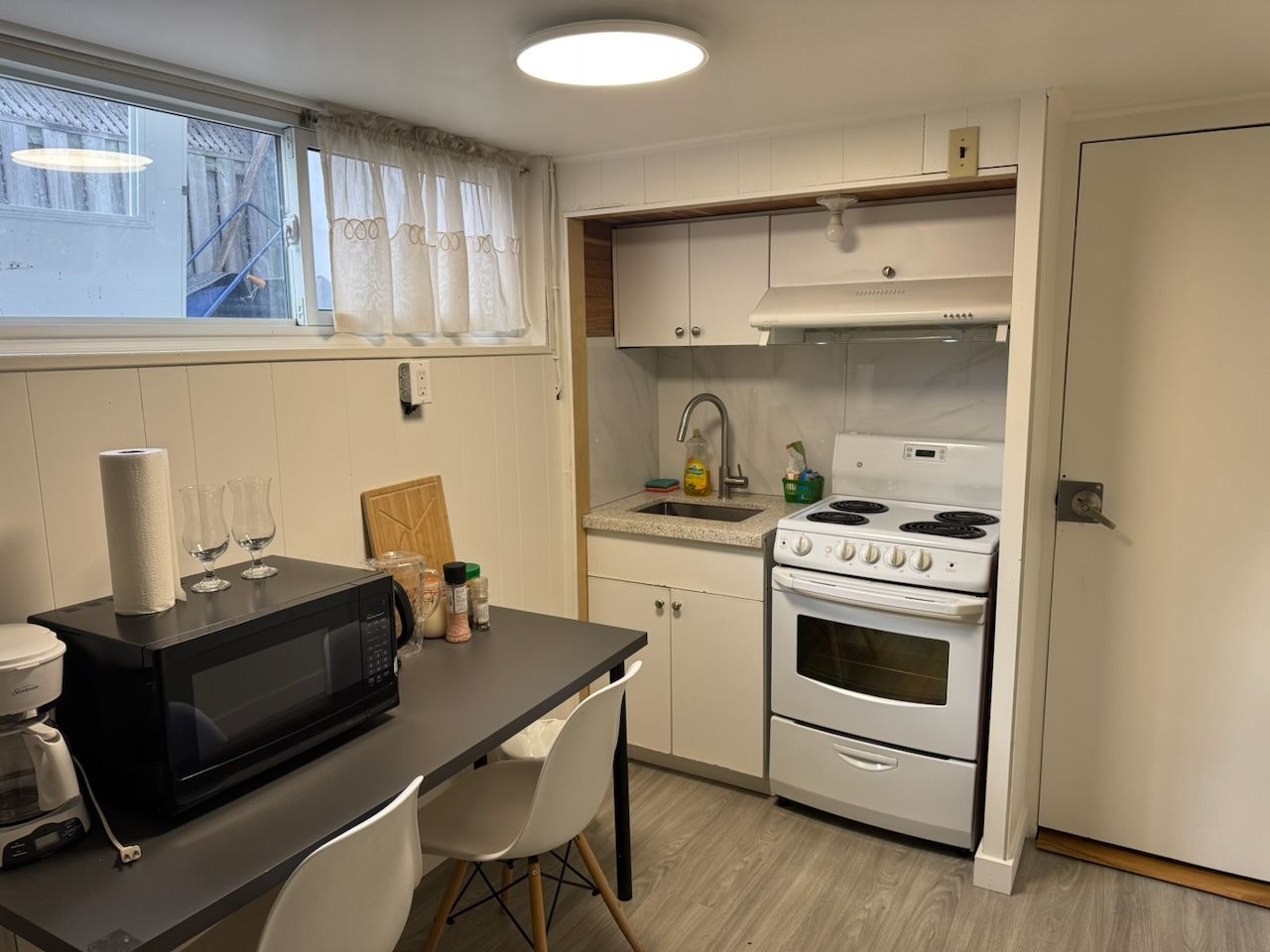 Une cuisine compacte équipée d’un four à micro-ondes, d’une cuisinière et d’un évier. On y trouve une fenêtre avec des rideaux, une chaise et une petite table sur laquelle sont posés un rouleau d’essuie-tout et un verre.