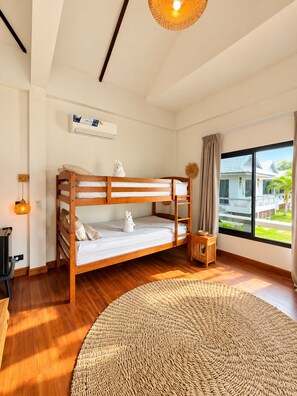 A bunk bed room with a woven rug, a small wooden side table, and a window with a view of a house. - Amana Beach Resort (Bophut)