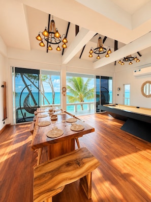 Um quarto espaçoso com uma mesa de jantar de madeira, mesa de bilhar e vista para o oceano através de grandes janelas.