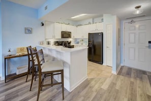 A modern kitchen with white cabinetry, a central island, stainless steel appliances, and a dining area with wooden chairs. - Pool Views,  2 Bed 2 Bath,Balcony, Lagoon Pool,Shuttle,The Palms of Destin (Destin)
