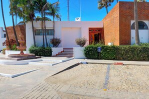 A building with a white facade, red brick accents, and a red door. There are palm trees and a fountain in the foreground. - Ocean View Condo -  Pools, Beach Access & More (Playas de Rosarito)