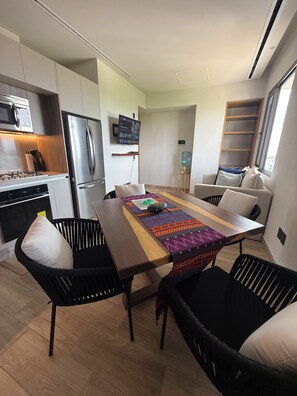 Una cocina moderna con microondas, estufa y refrigerador integrados. Hay una zona de comedor con una mesa de madera, sillas negras y un camino de mesa colorido. Se ve un sofá y un dispensador de agua al fondo.
