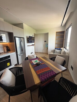 Una cocina moderna con electrodomésticos de acero inoxidable, un comedor con una mesa larga y sillas negras, y una sala de estar con un sofá y un ventanal.