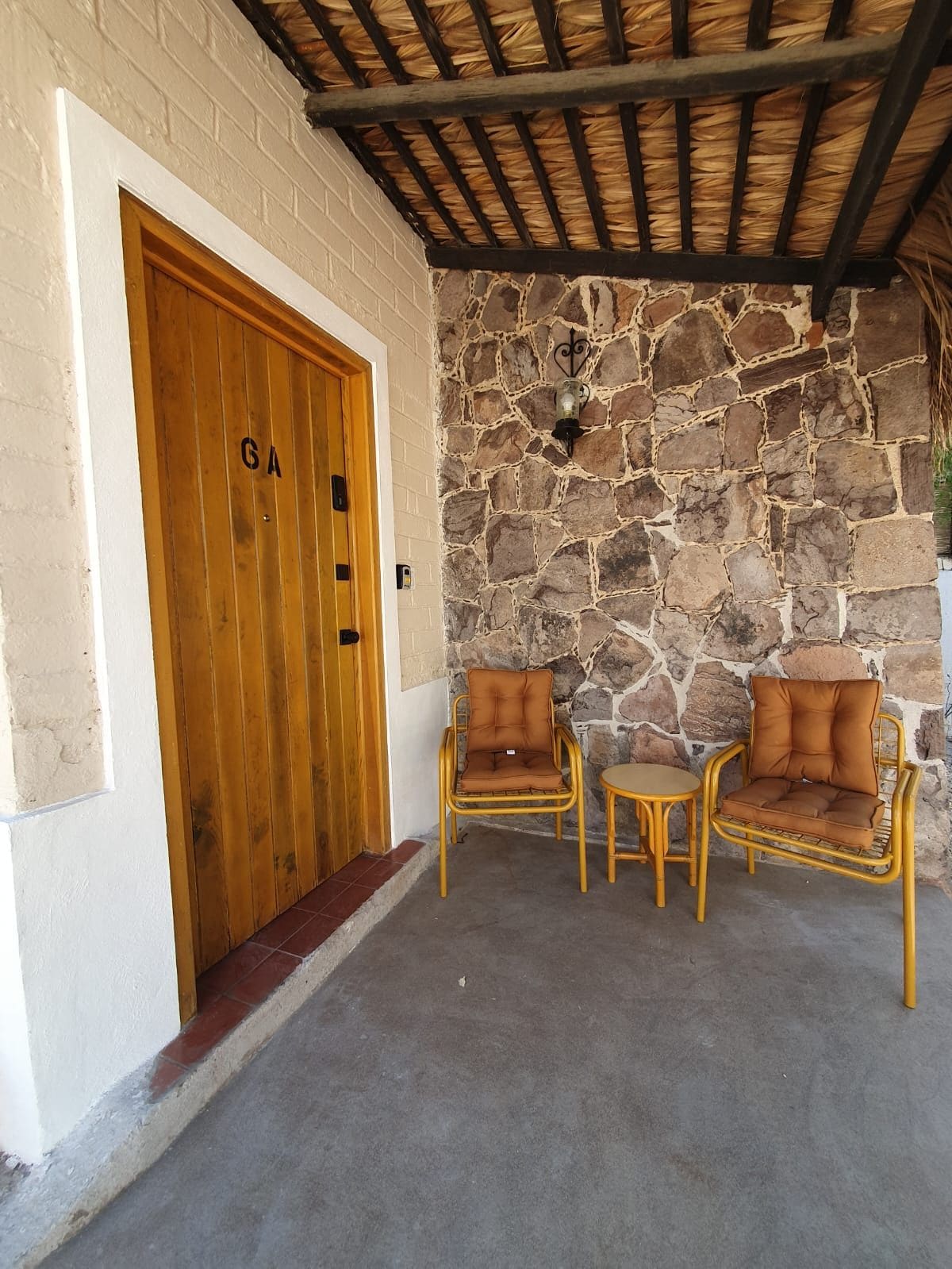 Un muro de piedra, una puerta de madera con un cartel que dice 'GA' y dos sillones de mimbre con cojines.