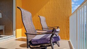 To flettede liggestole med puder på en balkon med en gul væg med struktur og udsigt til havet.