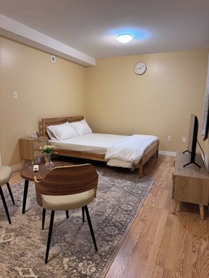 A hotel room with a bed, bedside table, chair, desk, and a clock on the wall. - Your Another Family Home (Beechville)