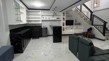 Un amplio salón con piso de mármol, un sofá, una cocina con armarios blancos y una escalera.