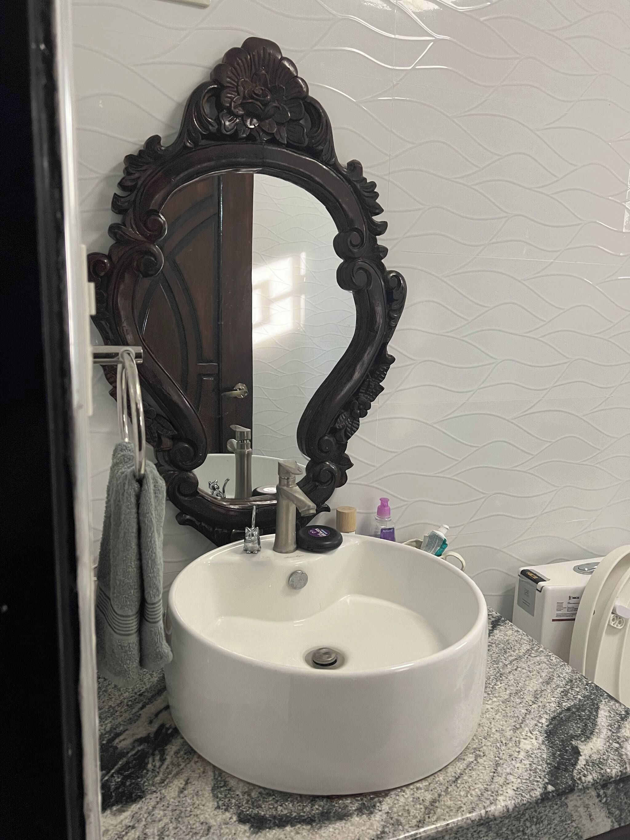 Une salle de bain avec un lavabo rond blanc, un grand miroir à cadre sombre et un comptoir en marbre.