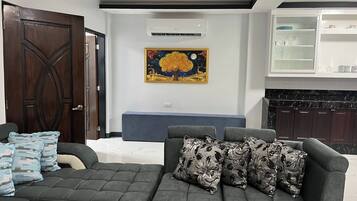 Un salon avec un canapé foncé, des coussins décoratifs, un climatiseur fixé au mur et un tableau.