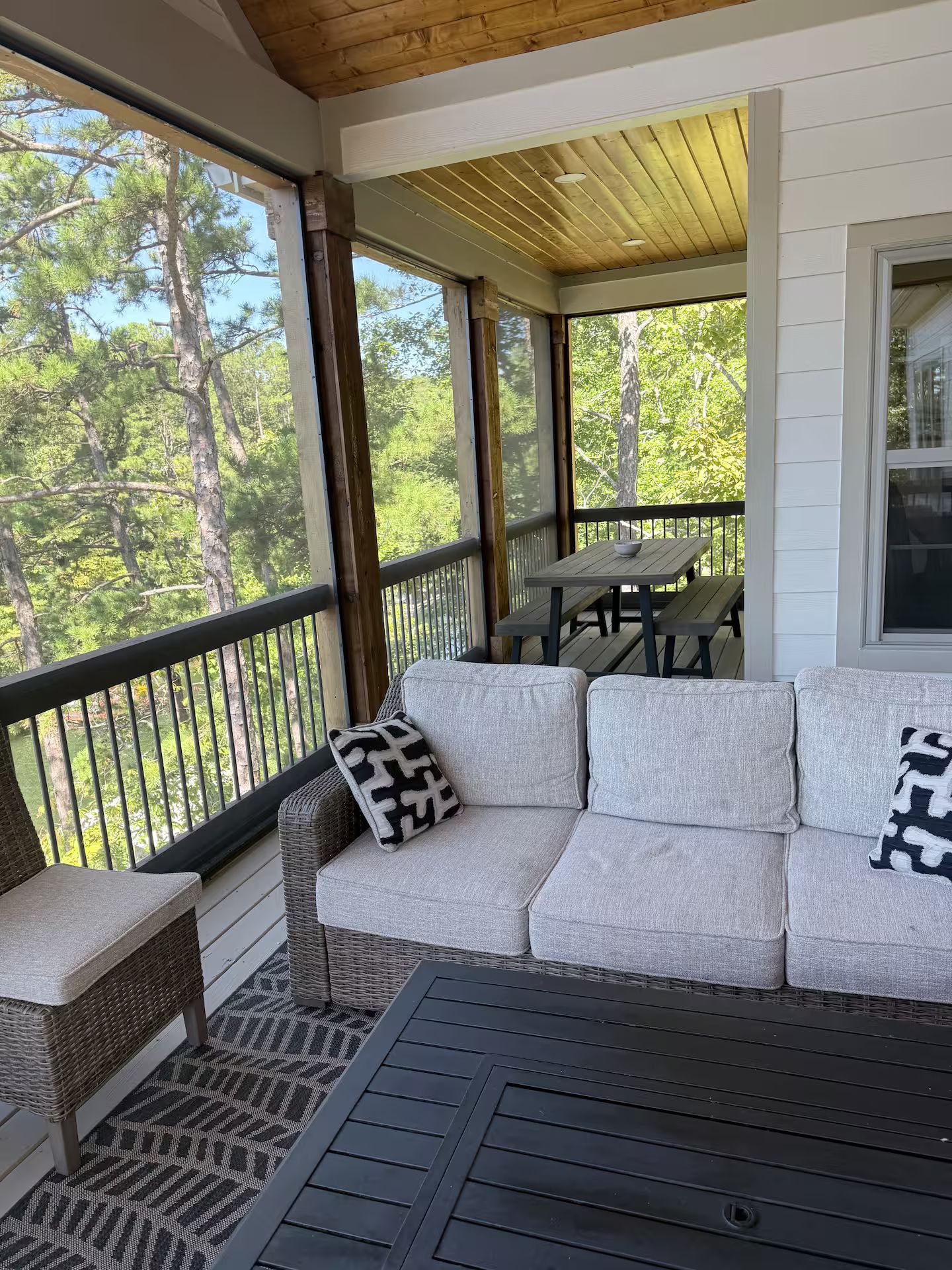 Eine überdachte Veranda mit einem Rattansofa, einer Bank und einem Tisch, mit Blick auf ein bewaldetes Gebiet.