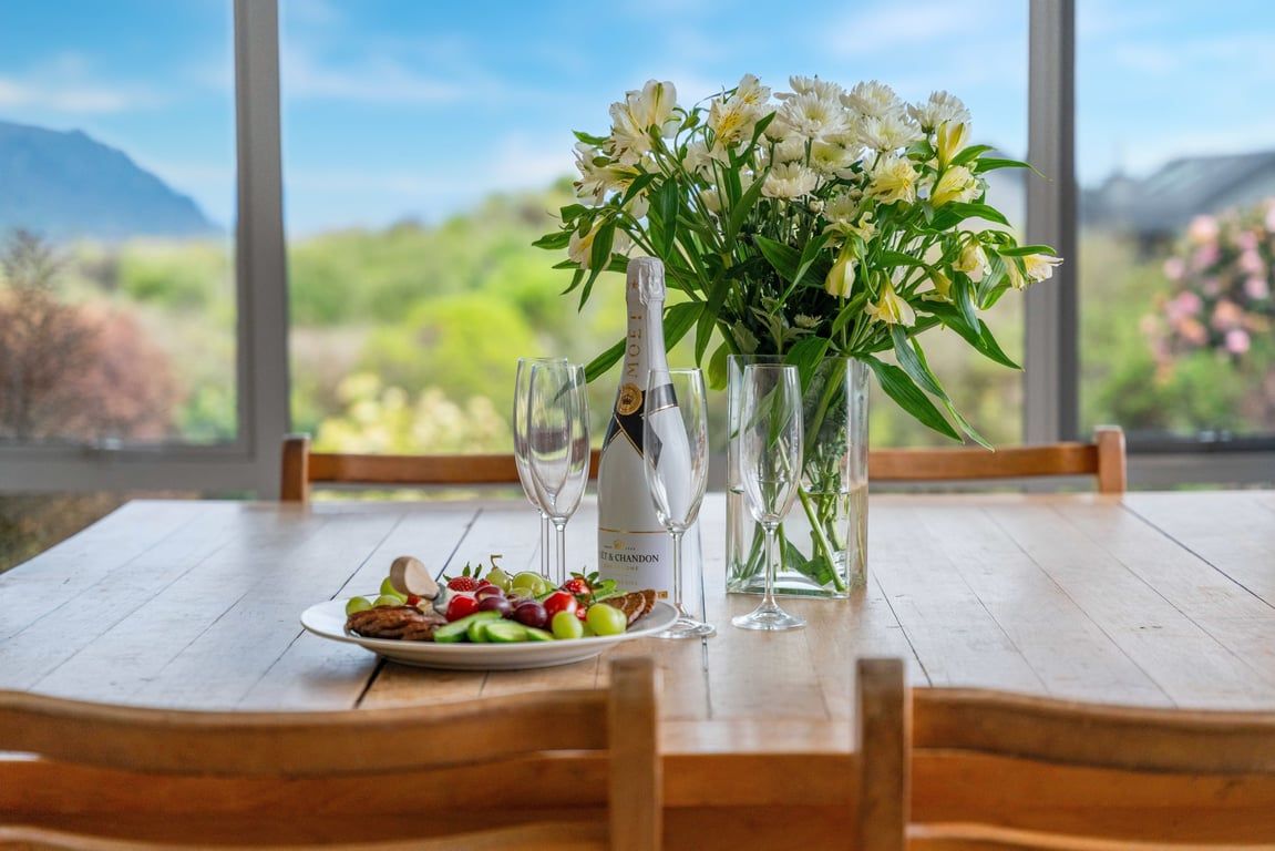 A dining table with a plate of food, two glasses, a bottle of champagne, and a vase of flowers.