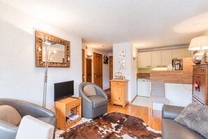 Una sala de estar con televisor, dos sofás, una mesita de madera, un tapiz de cuero de vaca y una cocina con armarios y estufa.