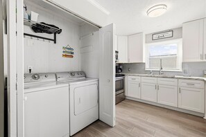 Un área de lavandería con lavadora y secadora, una cocina con armarios blancos y un ventanal, y un letrero en la pared.