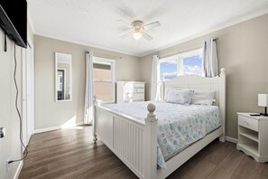 Quarto com cama, mesa de cabeceira, cômoda, ventilador de teto e janela com cortinas.
