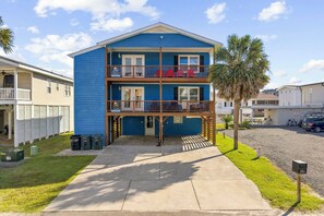 A two-story building with a balcony on the upper level, a covered entrance, and a parking area with a palm tree. - Family Tides: Unit 2 - Comfy 3BR Unit w/ Private Balcony (Walk to Beach) (North Myrtle Beach)