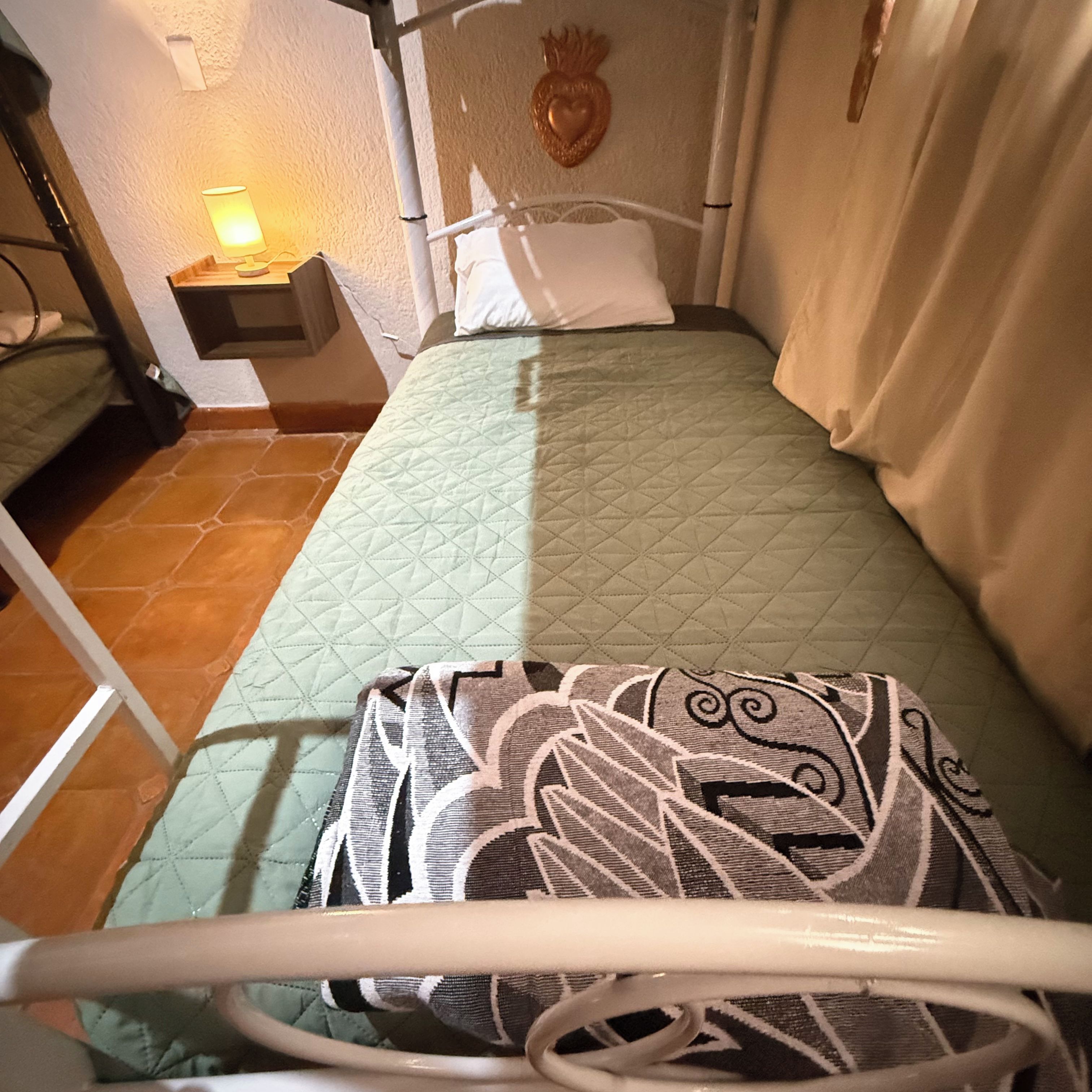 Una cama con colcha verde acolchada, un cojín estampado y una lámpara de noche sobre una mesita.