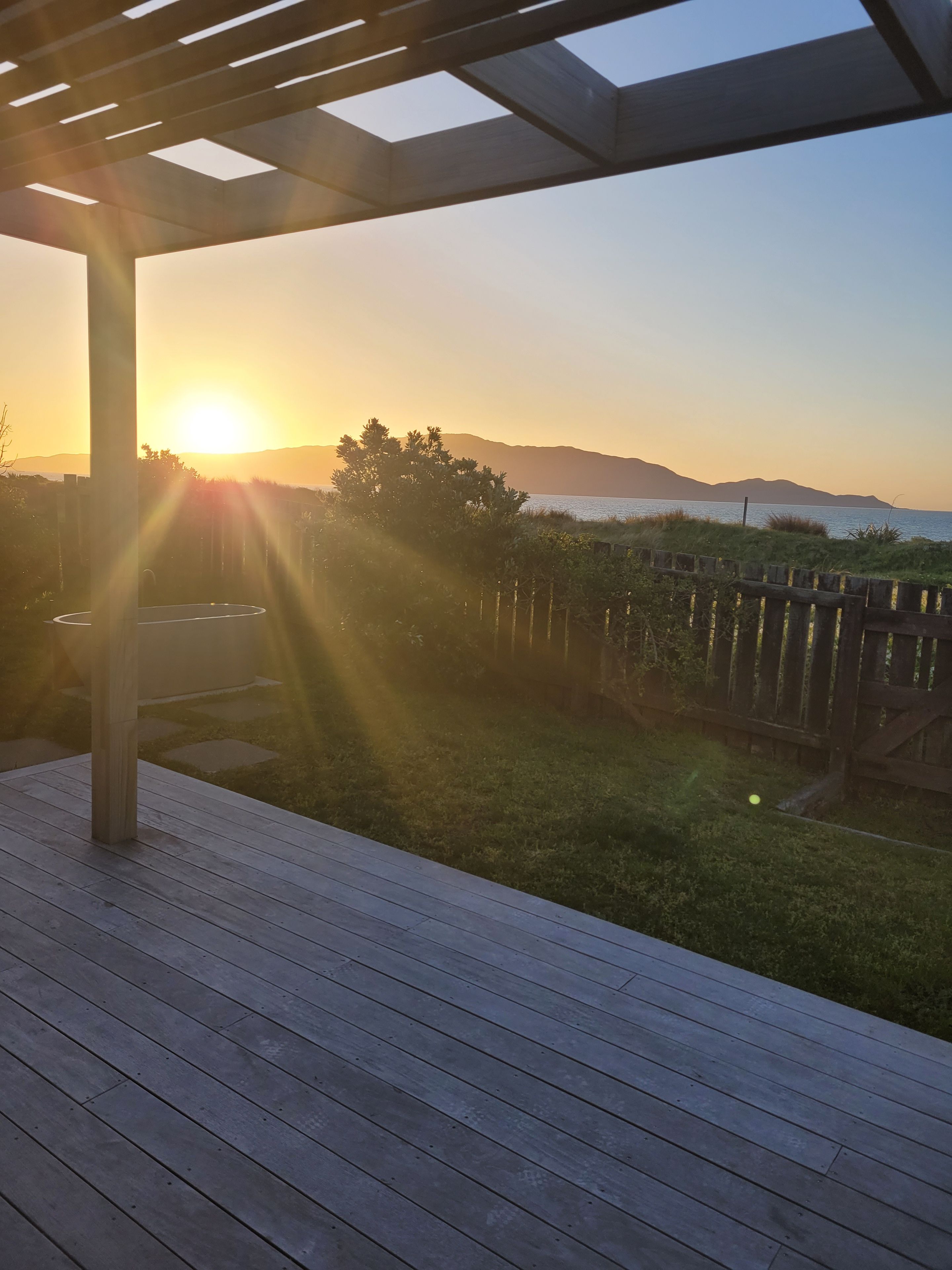 A wooden deck with a pergola structure, overlooking a grassy area with a wooden fence and a body of water in the distance.