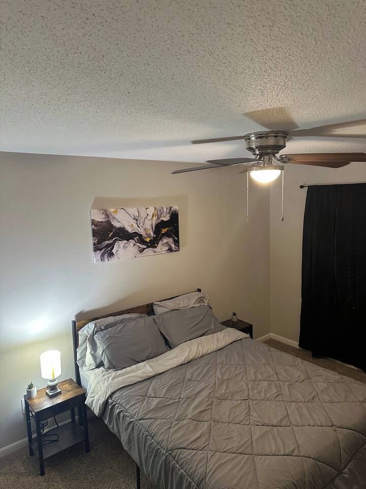 Une chambre à coucher comprenant un lit, une table de chevet, une lampe, un ventilateur de plafond et une œuvre d’art accrochée au mur.