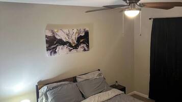 Un dormitorio con cama, mesita de noche, lámpara, ventilador de techo y una obra de arte en la pared.