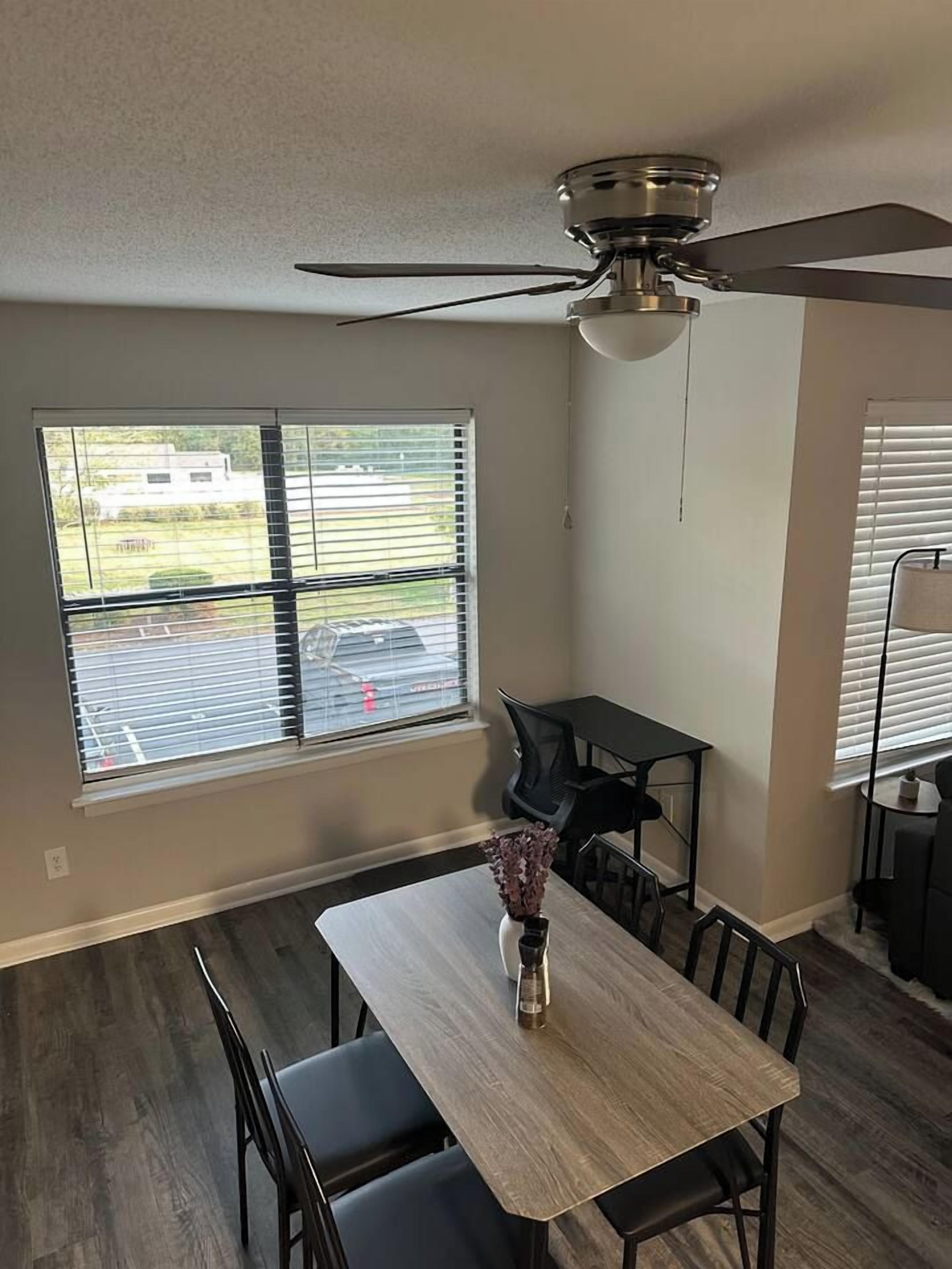 A dining area with a table, chairs, a ceiling fan, and windows with blinds.