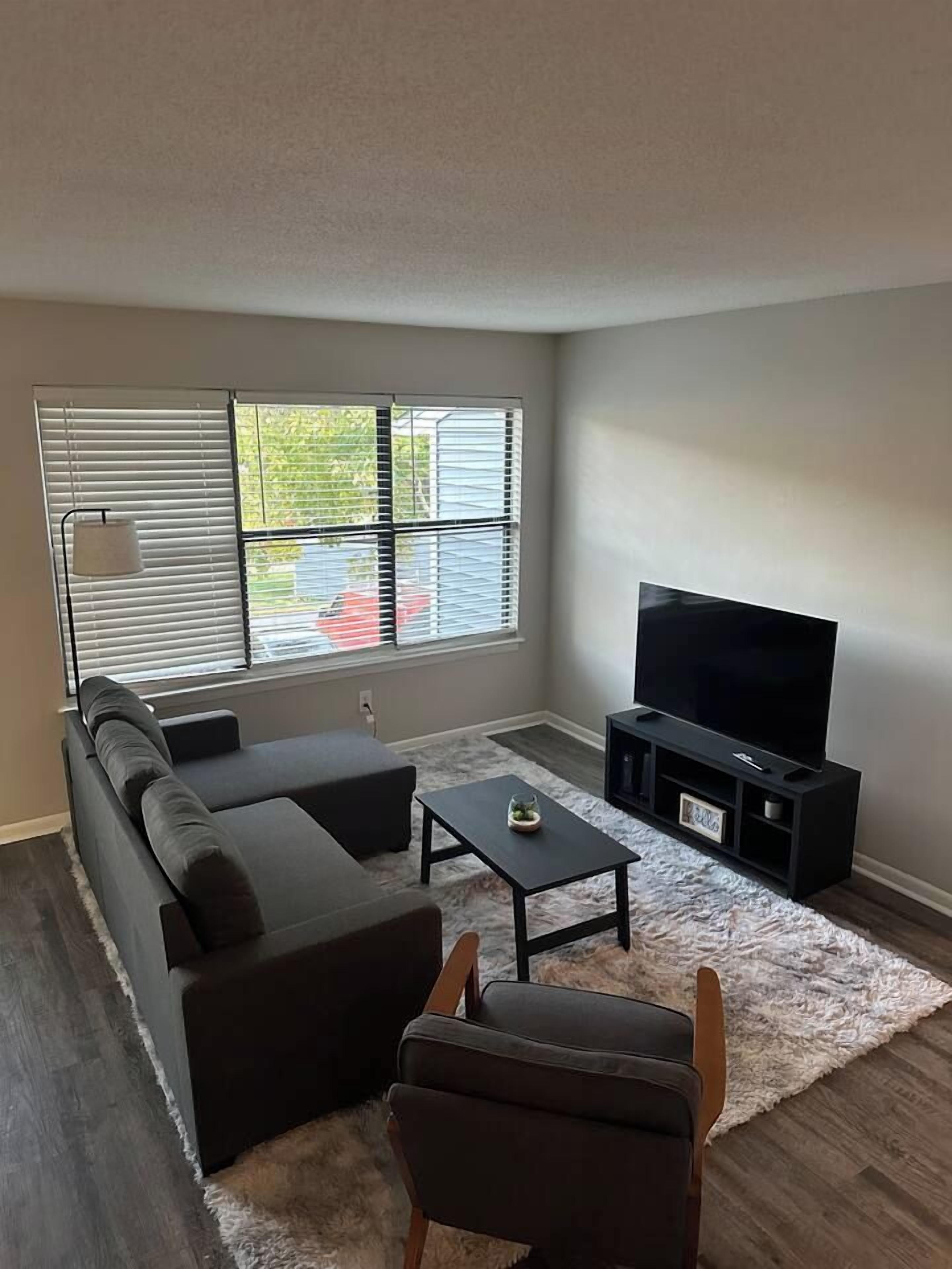 A living room with a sectional sofa, a flat-screen TV on a stand, a coffee table, and a window with blinds.