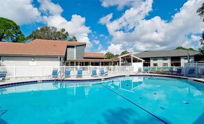 A swimming pool with lounge chairs, a covered seating area, and a building with a tiled roof. - Lakefront Shorewalk Palms Condo Easy Does It (Bradenton)