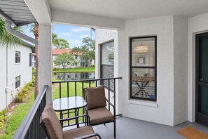 A balcony with a glass table and two chairs, a view of a pond, and a building with a red roof in the background. - Lakefront Shorewalk Palms Condo Easy Does It (Bradenton)