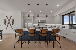 A modern kitchen with four bar stools, a stainless steel refrigerator, and pendant lighting. - New Luxury 4 bed | Hot Tub w/ Teton View | Targhee | AC (Driggs)