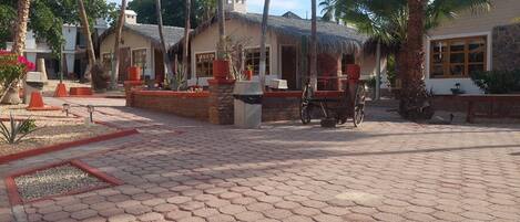 A courtyard with hexagonal paving, palm trees, and buildings with thatched roofs.