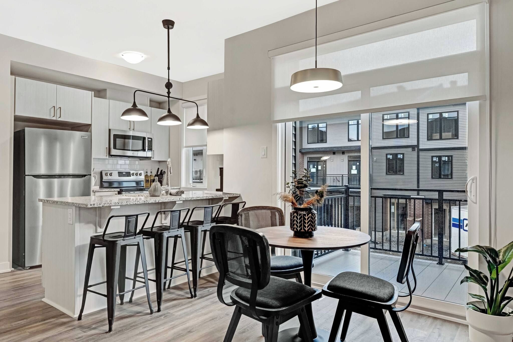 A modern kitchen with stainless steel appliances, a dining area with black chairs, and a view of a building through large windows.