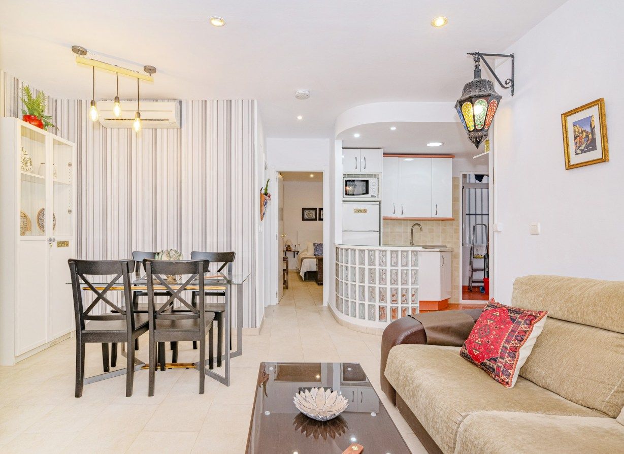 A modern living room with a sofa, a dining table with chairs, a kitchen area with cabinets and a microwave, and a hallway leading to another room.