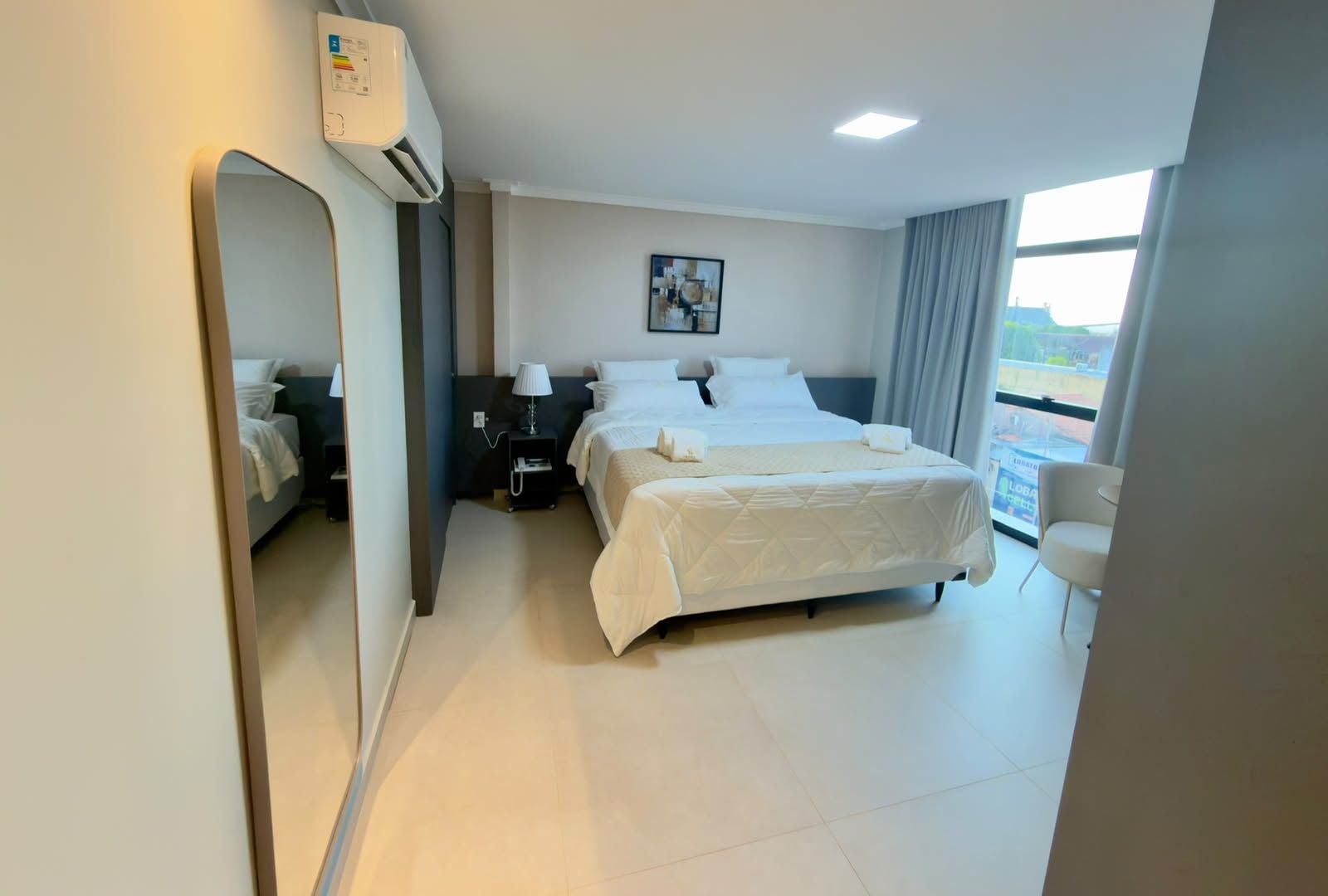 Habitación de hotel con una cama grande, una mesita de noche, una lámpara, un espejo y una ventana con cortinas.
