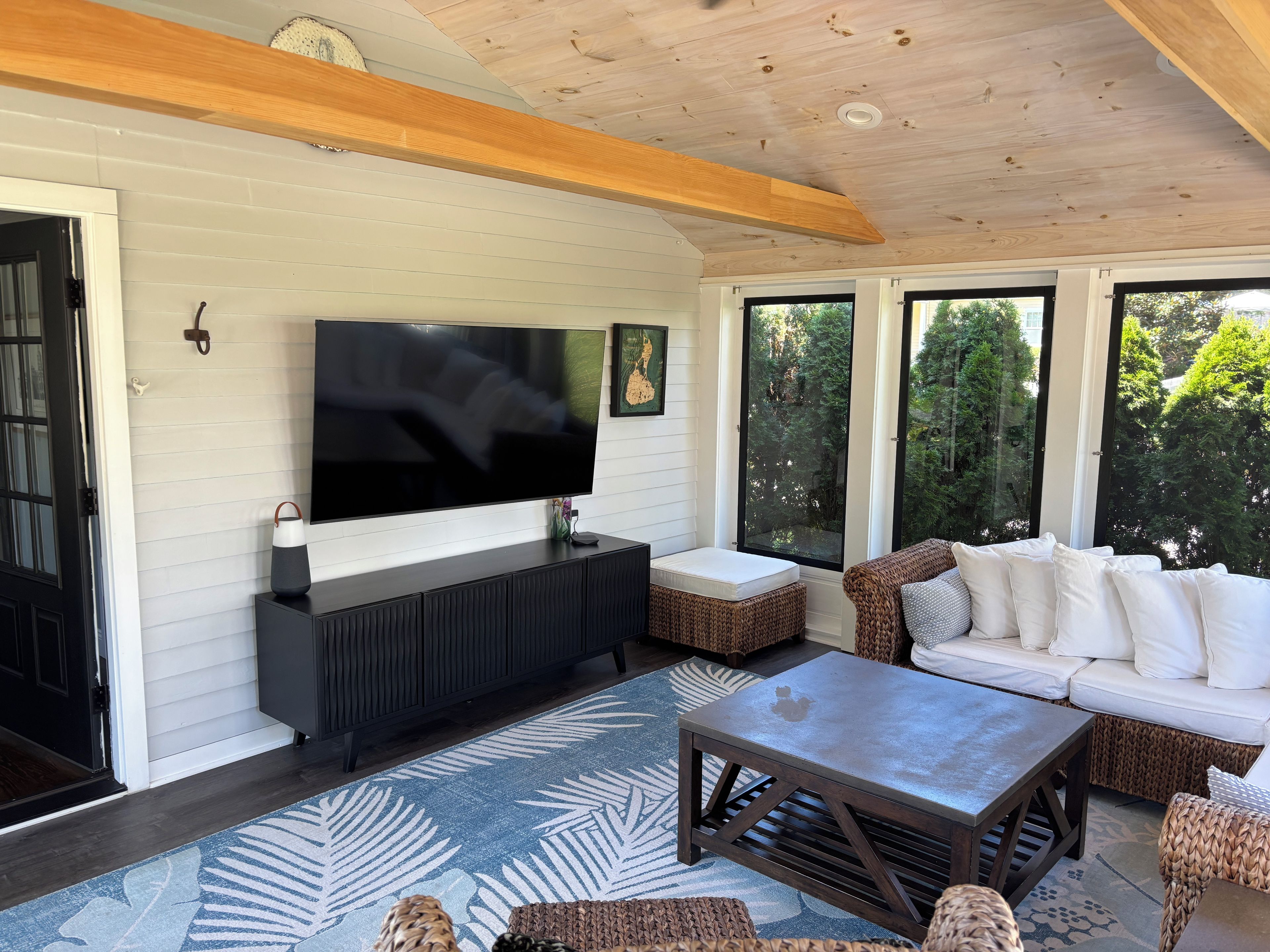 A modern living room with a flat-screen TV, dark wood furniture, and large windows offering a view of greenery.