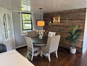 Et spiseområde med et rundt bord, fire stole, en vase med blomster, en hængende lampe og en trævæg med billedramme.