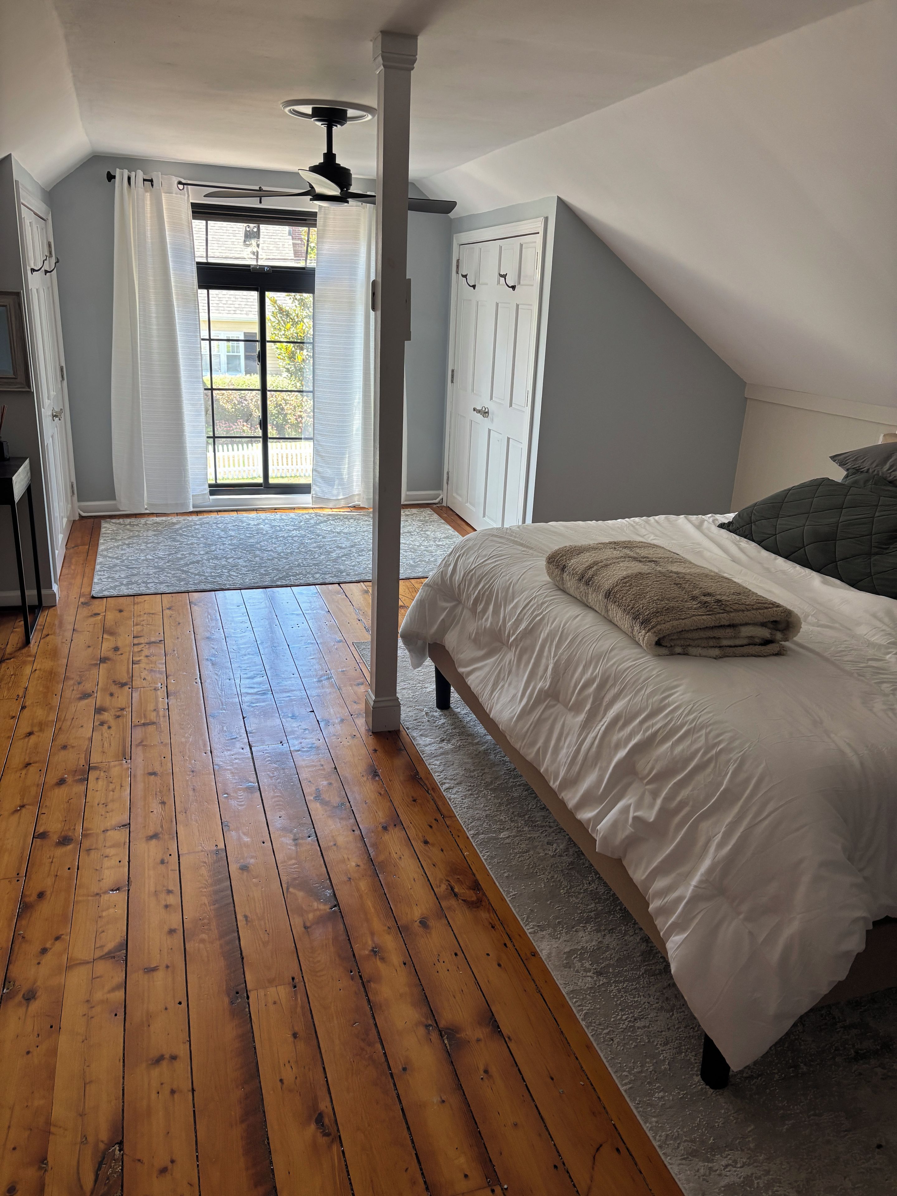 Una camera da letto con pavimento in legno, un letto con biancheria bianca, una ventola a soffitto e vista sull'esterno attraverso una porta.
