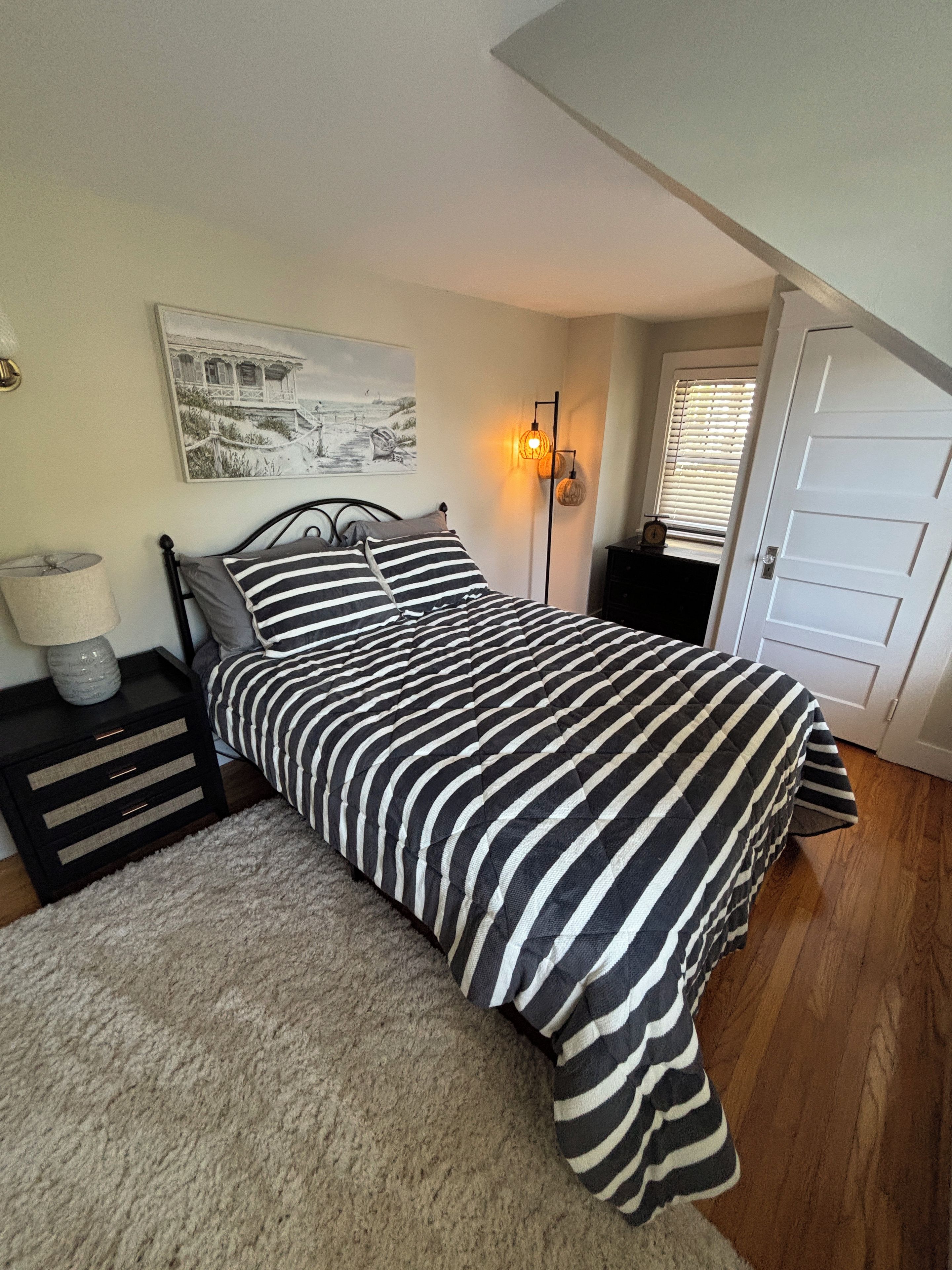 A bedroom with a bed, a nightstand, a lamp, a wall clock, and a door.