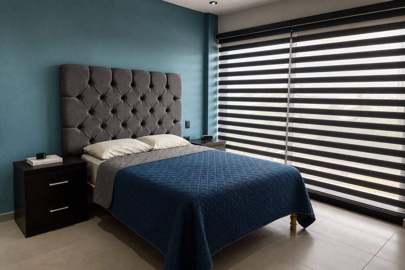 A bedroom with a tufted headboard, a bed with a blue quilt, a nightstand with a book, and a window with horizontal blinds.