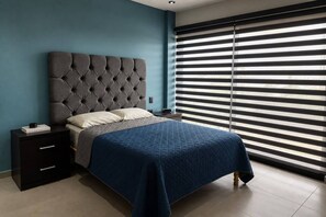 Una camera da letto con una testiera trapuntata, un letto con copriletto blu, un comodino con un libro e una finestra con persiane orizzontali.