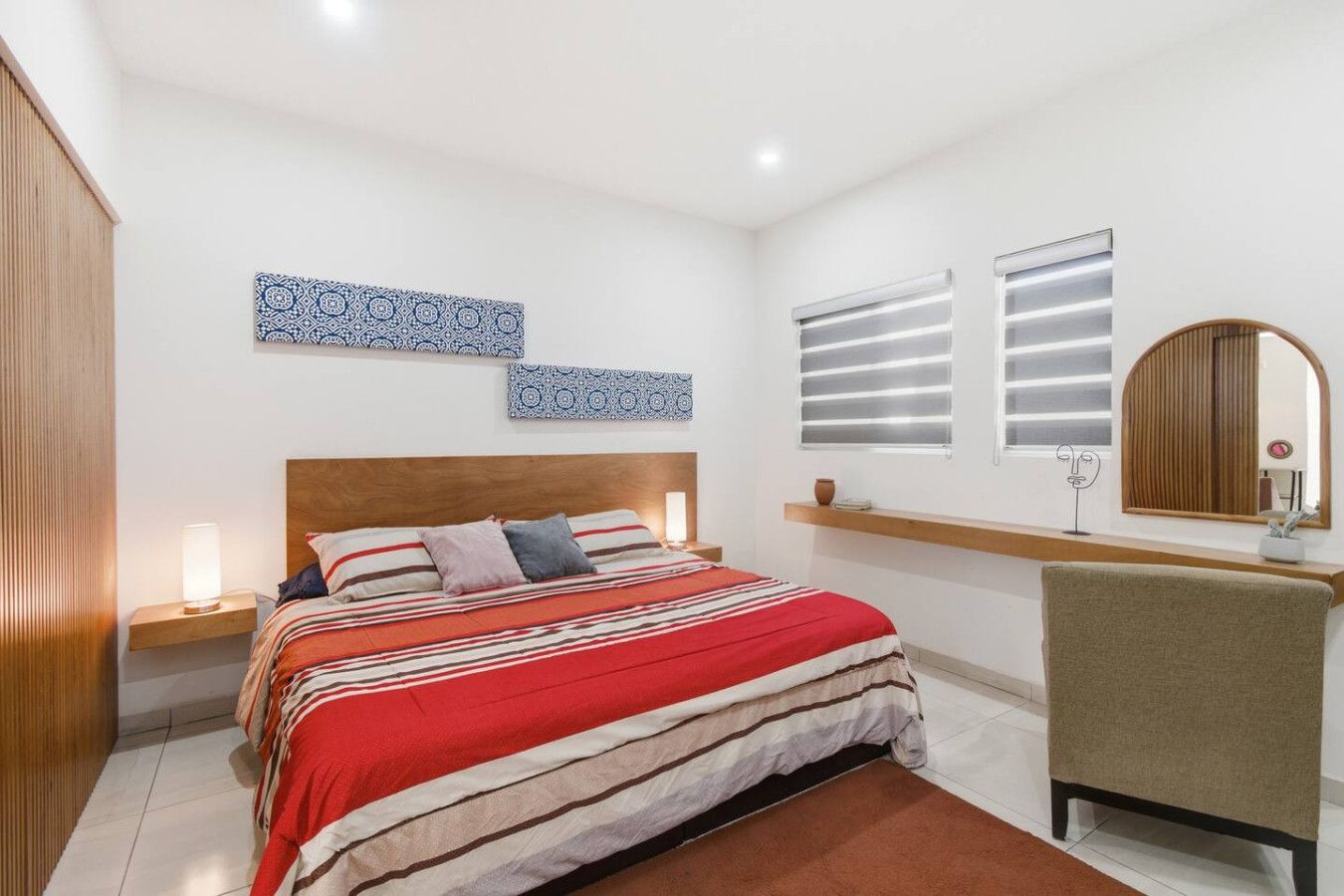 A bedroom with a bed, bedside tables, a chair, a mirror, and wall art.