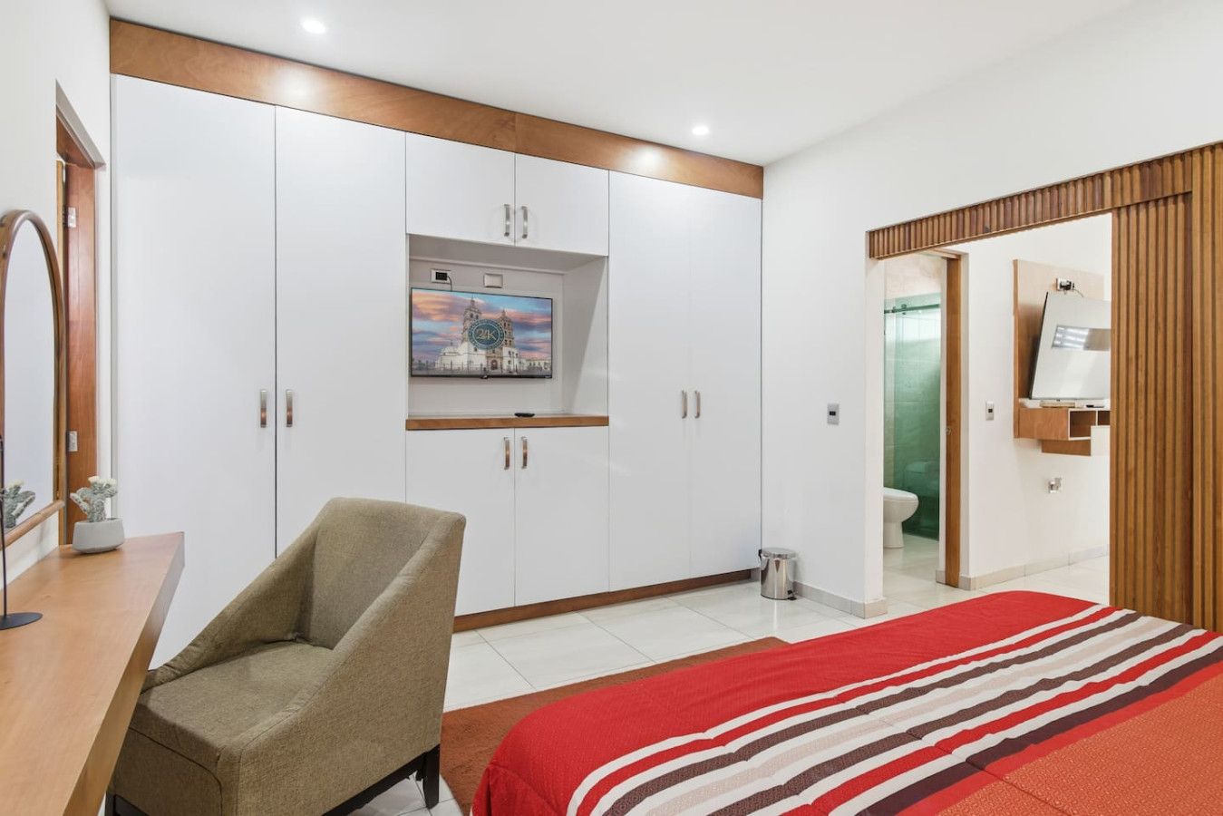 A bedroom with a bed, a chair, a wardrobe, a TV, and a bathroom visible through an open door.
