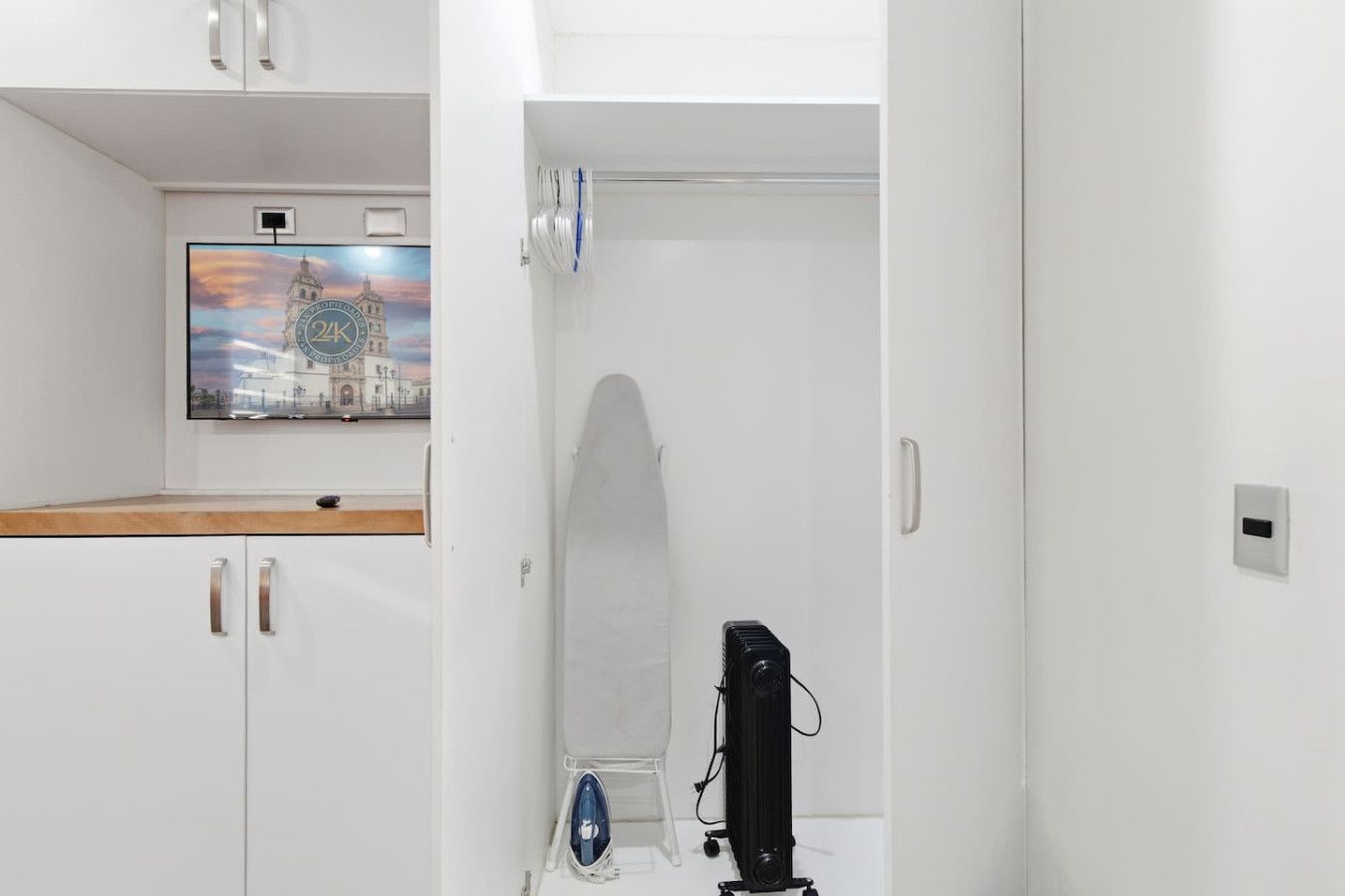 A room with a white wardrobe, a flat-screen TV mounted on the wall, and a surfboard leaning against the wall.