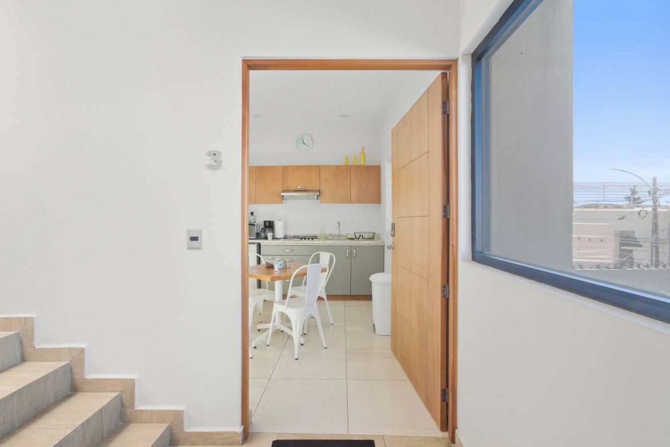 A modern kitchen with wooden cabinets, a dining area with white chairs, and a large window offering a view of the city.