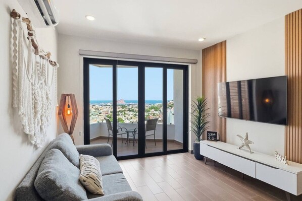 Een moderne woonkamer met een grijze bank, een flatscreen televisie en een balkon met uitzicht op de stad.