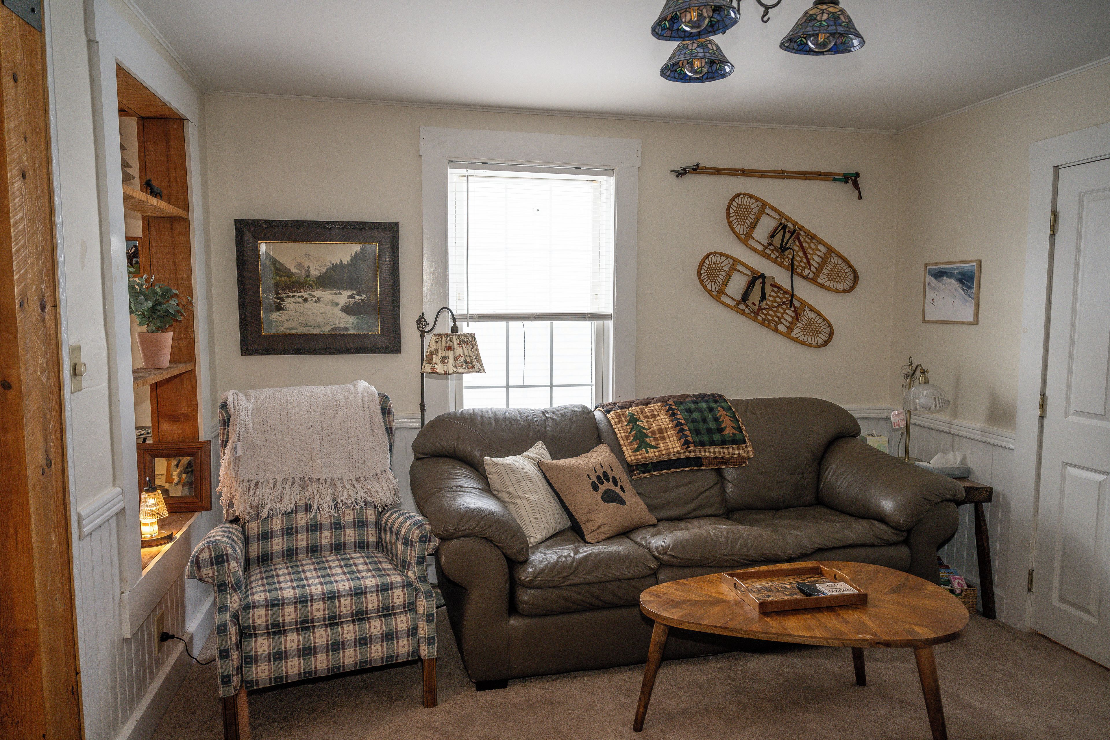 A living room with a brown leather sofa, a plaid armchair, a wooden coffee table, and a window with blinds.