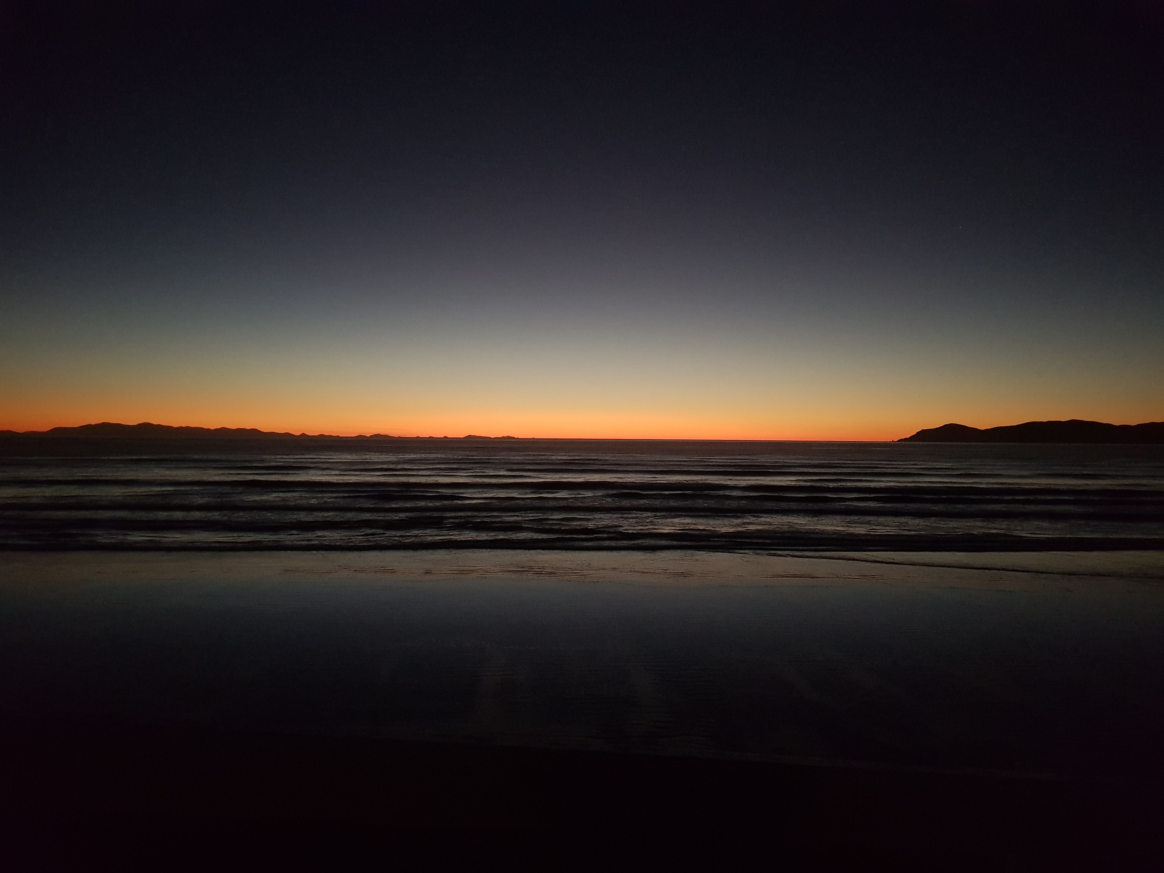 Una spiaggia al tramonto con onde e montagne in lontananza.
