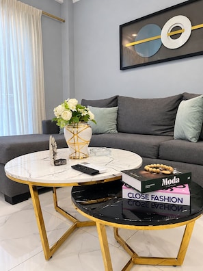 En moderne stue med en grå sofa, et rundt sofabord med gyldne ben, en vase med hvide blomster og et indrammet kunstværk på væggen.