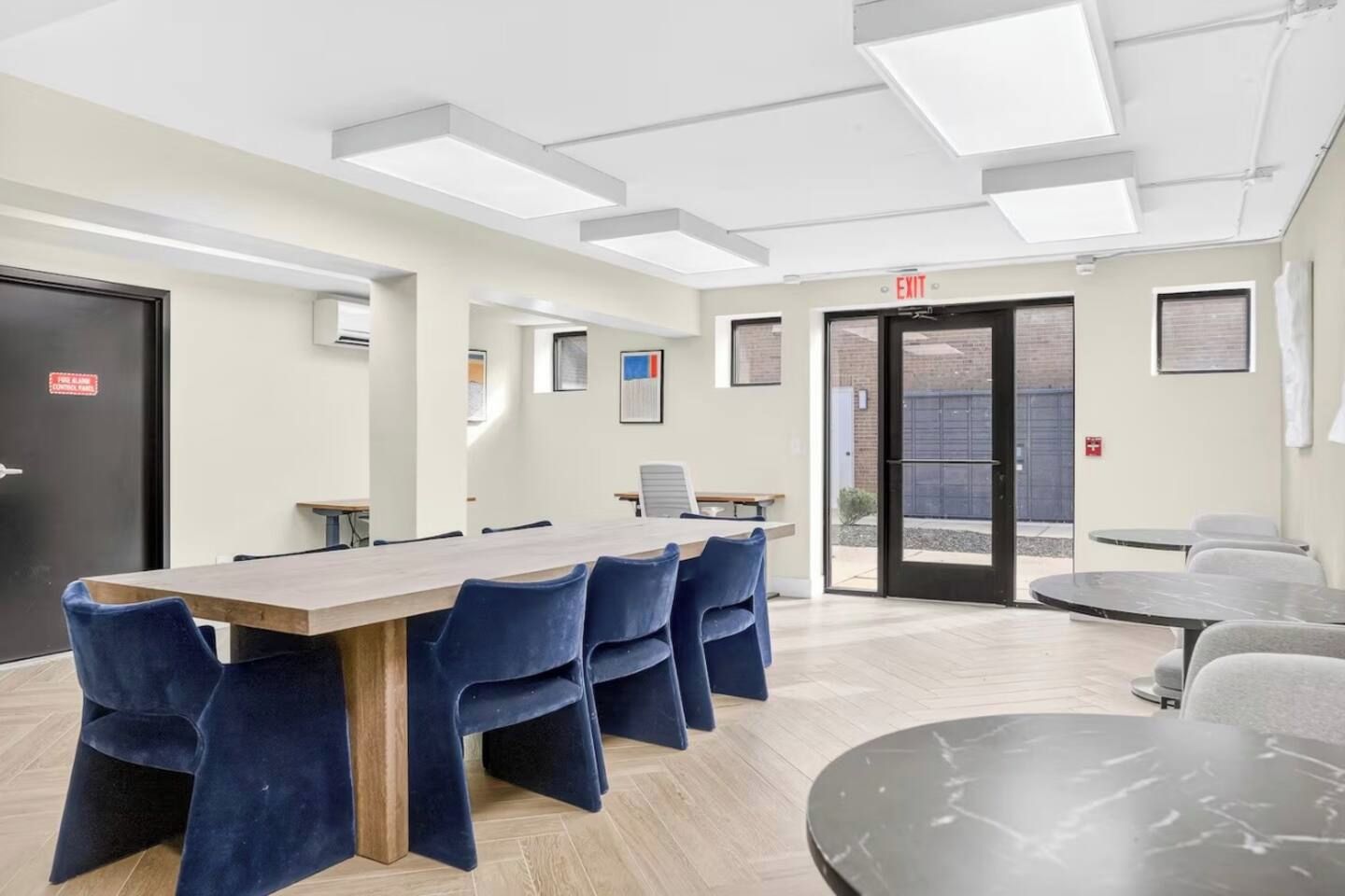 A conference room with a long table and blue chairs, a door with an exit sign, and a view of the outside.